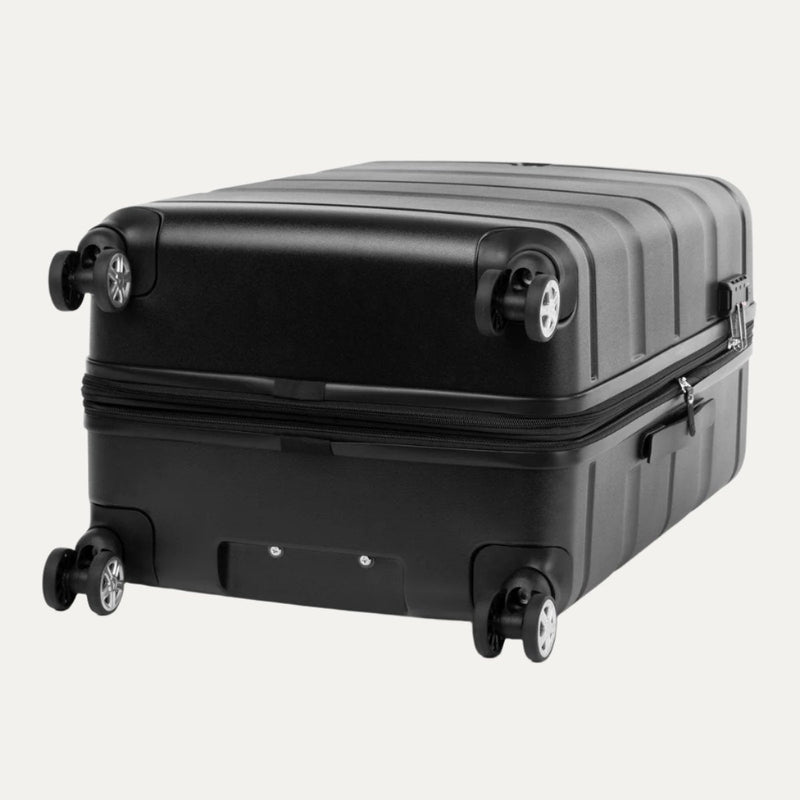 Black hardside spinner suitcase shown lying flat on its back, displaying the bottom view with four dual spinner wheels, a central rectangular recessed area, and the zipper closure running around the middle.
